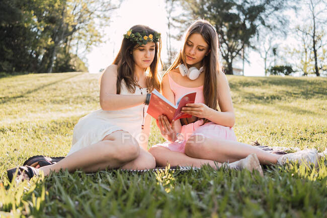 Подружки в сарафанах діляться підручником, сидячи на лузі в сонячному парку в спині освітленому — стокове фото