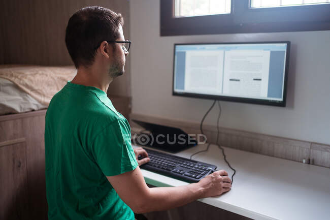 Боковой вид блогера-мужчины в очках, назидающего текст на мониторе, пока он сидит на клавиатуре в домашней комнате — стоковое фото
