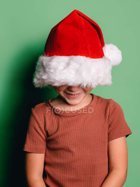 Irreconhecível feliz menino cobrindo rosto com vermelho Santa chapéu de pé contra fundo verde — Fotografia de Stock