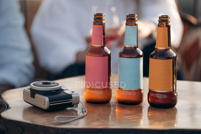 Crop anonymous friends sitting at table with glass bottles of beer near instant photo camera in bar — Stock Photo
