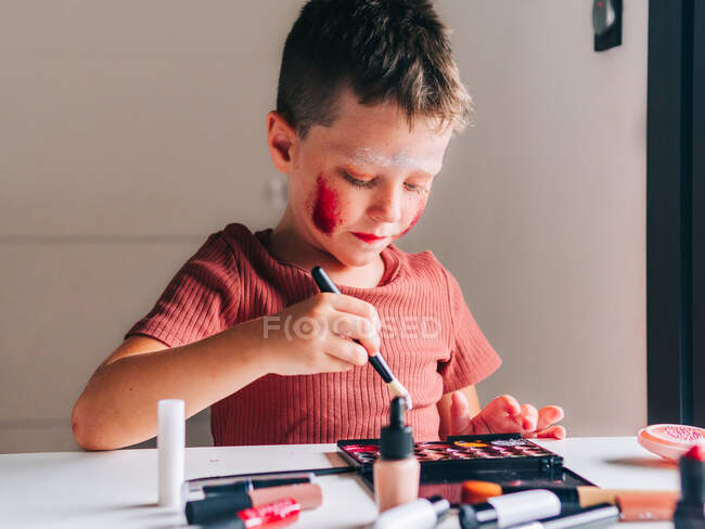 Очаровательный ребенок с аппликатором для макияжа смотрит на стол с палитрой теней для век — стоковое фото