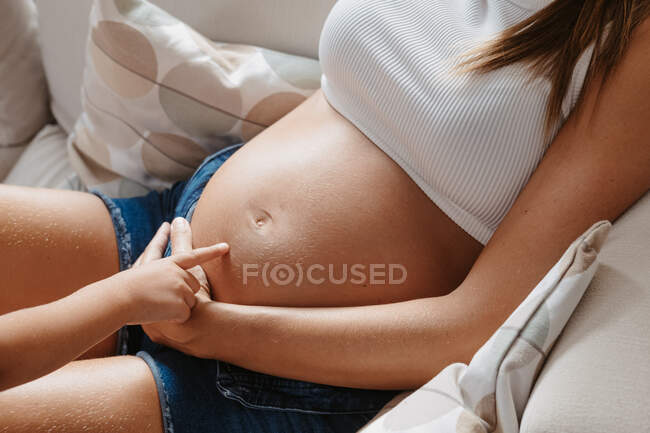 Zugeschnittenes, unkenntlich gemachtes Kind streichelt Bauch der werdenden Mutter auf der heimischen Couch — Stockfoto