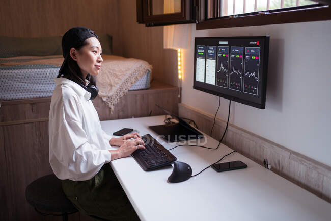 Vue latérale de concentré asiatique femme travaillant sur ordinateur avec des graphiques montrant la dynamique des changements de valeur de crypto-monnaie à un lieu de travail pratique — Photo de stock