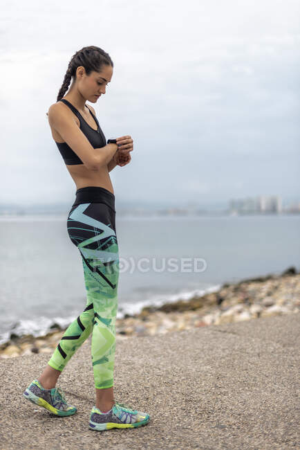 Side view of fit female runner checking pulse on fitness tracker while standing on embankment during cardio training — Stock Photo