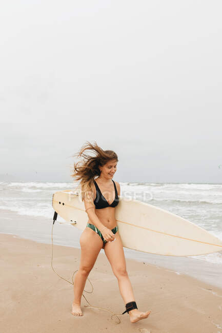Joyeux jeune sportive en maillot de bain avec planche de surf regardant loin sur la côte sablonneuse contre l'océan orageux — Photo de stock