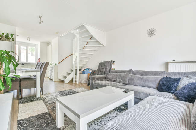 Innenraum eines hellen Zimmers mit Esstisch und Stühlen gegen graues Sofa mit Kissen und Teppich tagsüber — Stockfoto