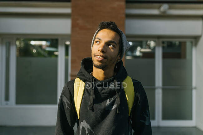 Handsome African American male hipster in hat and hoodie smiling and looking away while standing in urban street on sunny day — Stock Photo