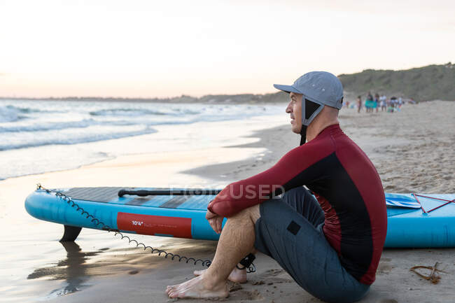 Vista laterale del ponderato surfista maschio in muta e cappello seduto guardando lontano con SUP bordo mentre si prepara a navigare sulla riva del mare — Foto stock