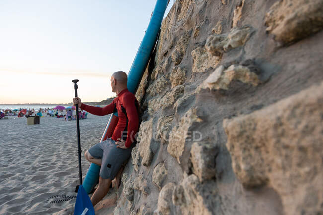 Side view of male surfer in wetsuit and hat standing looking away with paddle and SUP board while preparing to surf on seashore leaning on stone wall — Stock Photo