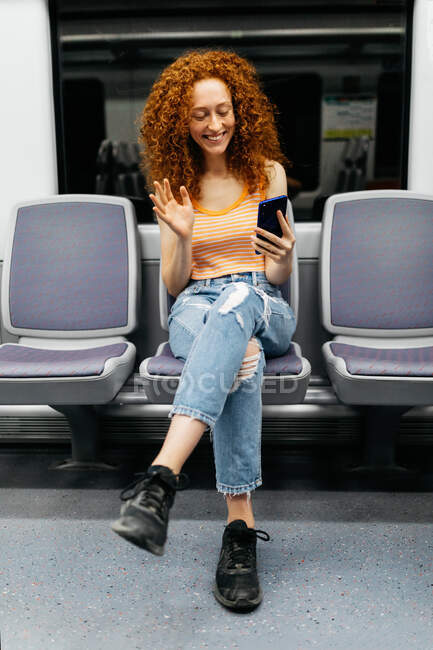 Friendly woman waving hand during video chat on cellphone while sitting with crossed legs on train — Stock Photo