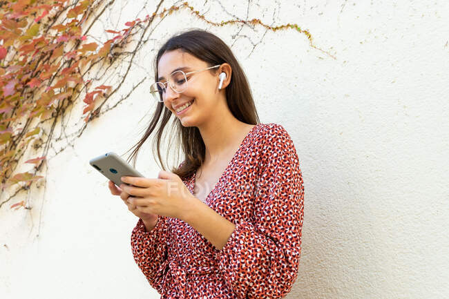 Cheerful female in casual clothes connecting wireless earphones with smartphone against light wall in daytime — Stock Photo