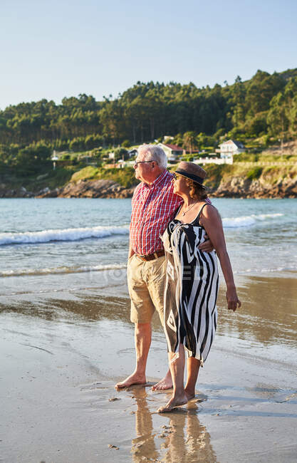 Улыбающаяся босиком пожилая пара в солнечных очках, стоящая на мокром песчаном пляже и наслаждающаяся солнечным днем — стоковое фото