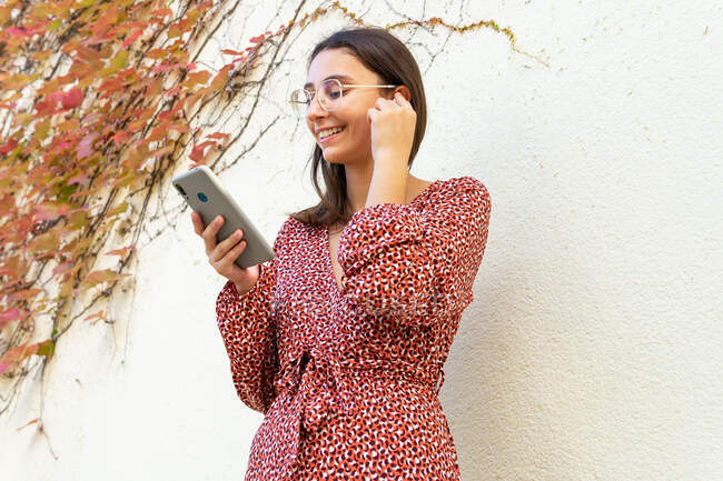 Весела жінка в повсякденному одязі, що з'єднує бездротові навушники зі смартфоном проти світлої стіни вдень — стокове фото