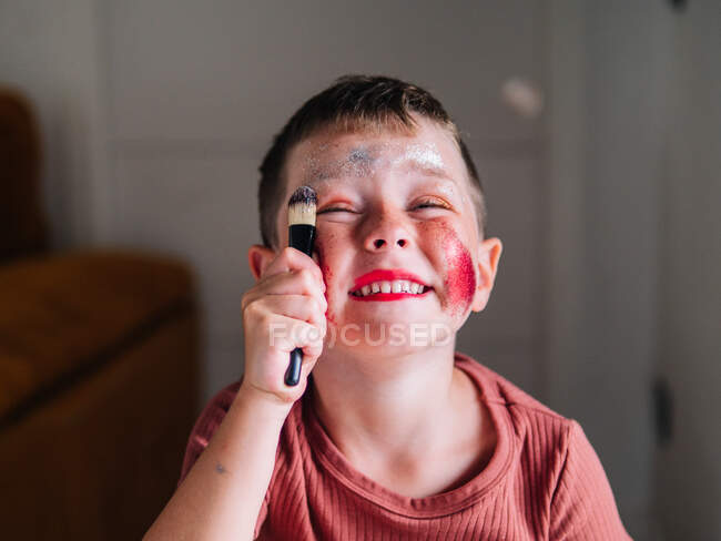 Niño encantador con aplicador de maquillaje mirando a la mesa con paleta de sombra de ojos - foto de stock