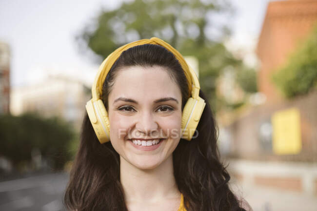 Positive young female listening to music in wireless headphones looking at camera while walking on the street — Stock Photo