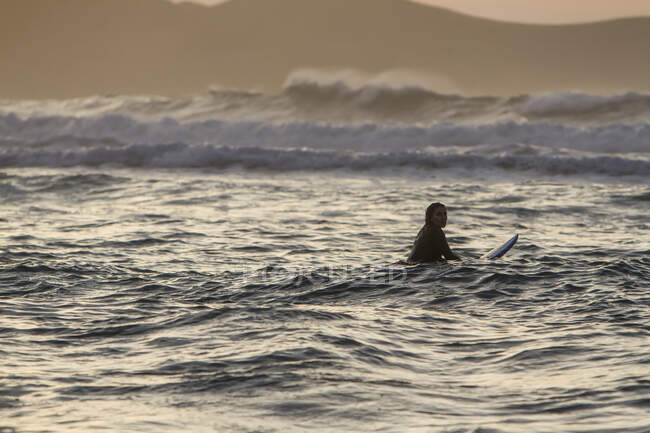 Vista trasera de una joven con tabla de surf en el mar al atardecer en la playa de Asturias, España - foto de stock