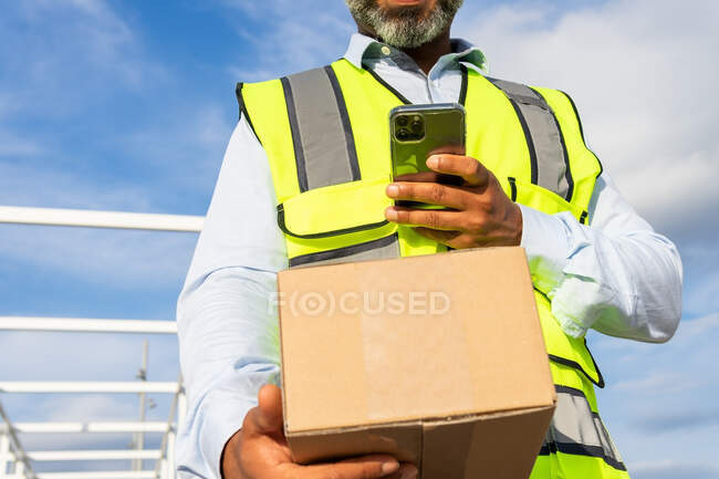 Trabajador masculino irreconocible recortado en uniforme que lleva el paquete mientras que usa el teléfono móvil en el trabajo - foto de stock