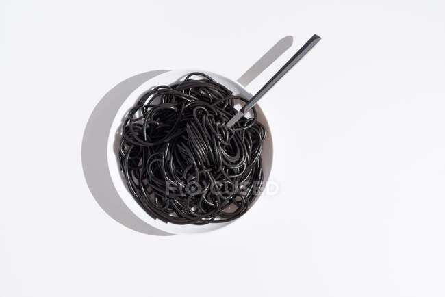 Vista superior de garfo inoxidável em tigela cheia de espaguete preto com tinta de choco na mesa branca em estúdio — Fotografia de Stock