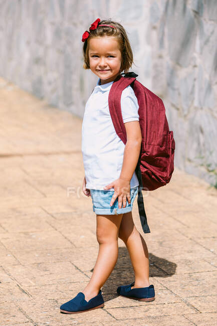 Vue latérale de l'écolier avec sac à dos sur la chaussée regardant la caméra en plein soleil — Photo de stock