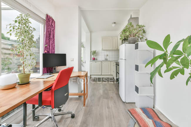 Table en bois avec ordinateur et plantes placées près de la fenêtre dans la pièce lumineuse près de la zone de cuisine dans l'appartement en journée — Photo de stock