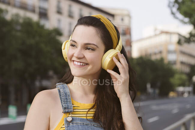 Mujer joven y positiva escuchando música en auriculares inalámbricos mirando hacia otro lado mientras camina por la calle - foto de stock