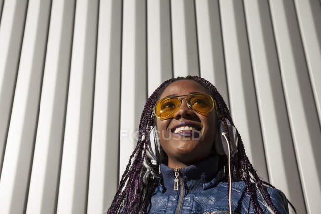 Von unten fröhliche junge Afroamerikanerin mit Afro-Zöpfen in blauer Jacke und stylischer Sonnenbrille, die Musik über Kopfhörer genießt, während sie im Sonnenlicht vor gestreifter Wand chillt — Stockfoto
