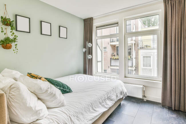 Dormitorio contemporáneo con almohadas en edredón entre plantas en maceta en casa con suelo de baldosas - foto de stock