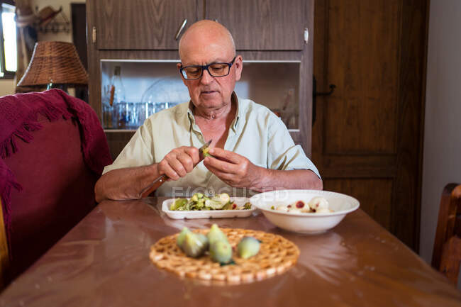 Senior male in eyeglasses with knife peeling green fig at table with disposable tray in house room — Stock Photo