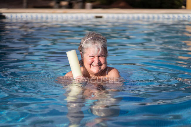 Femme âgée joyeuse avec des cheveux gris nageant dans la piscine avec une nouille aqua et souriant vivement à la caméra — Photo de stock
