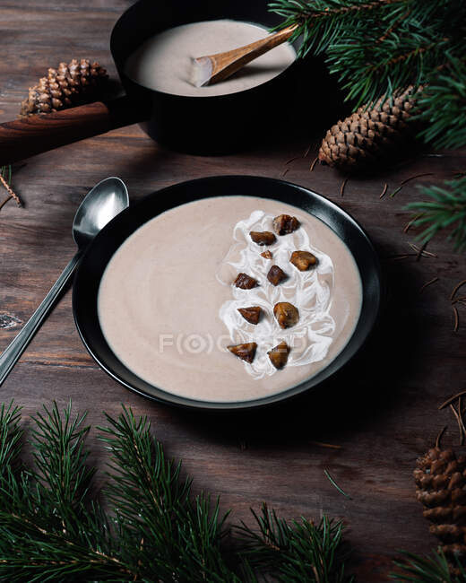From above of black bowls with cream soup of chestnuts placed on wooden table with green branches of fir with cones — Stock Photo