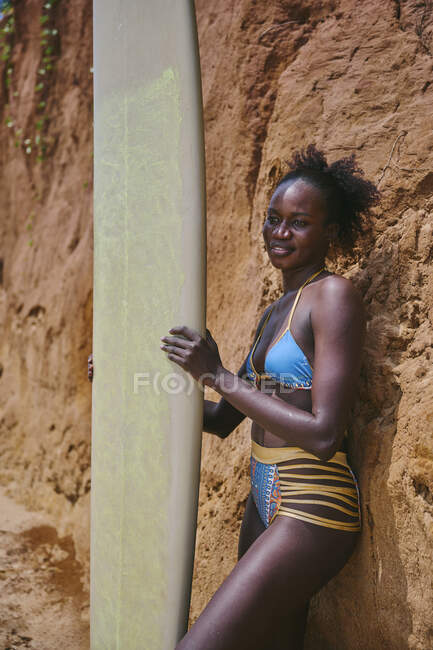 Side view of African American female athlete looking at away with surfboard from an area of the beach and in front of a clay rock — Stock Photo