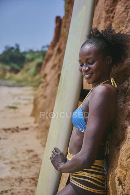 Vue latérale d'une athlète afro-américaine aux yeux fermés avec planche de surf depuis une zone de la plage et devant un rocher argileux — Photo de stock