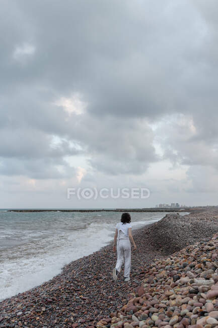 Vista posterior de una mujer anónima paseando por la orilla de guijarros contra el océano espumoso bajo nubes esponjosas al atardecer - foto de stock