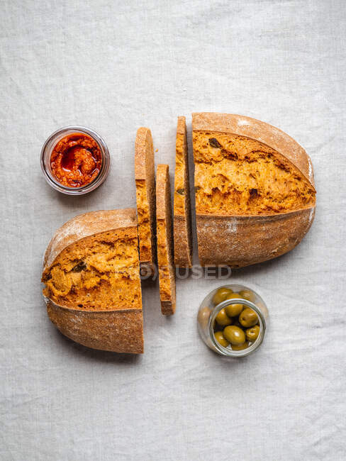 Top view of cut loaf of bread placed on table near red pesto sauce and jar of green olives — Stock Photo