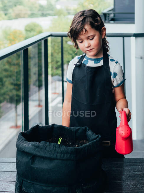 Маленька дівчинка з темним волоссям в фартусі стоїть і поливає рослину з горщиками на балконі вдень — стокове фото