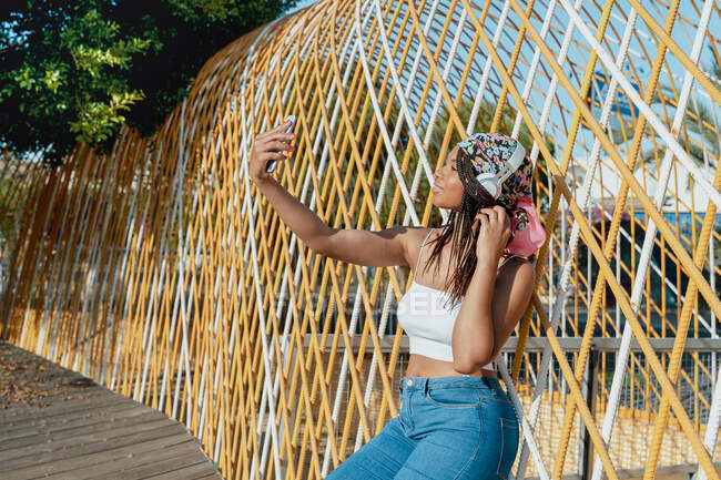 Giovane donna etnica positiva con cellulare che si fa un selfie mentre ascolta musica da cuffie wireless mentre ride in città — Foto stock