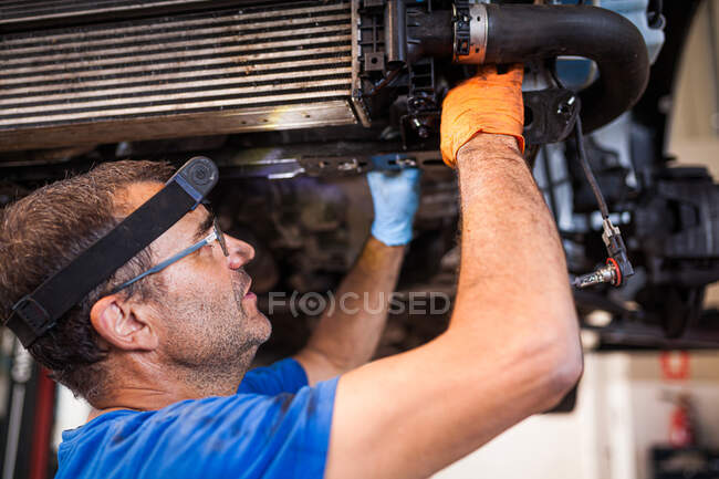 Ocupado maduro mecánico masculino en cabeza antorcha reparación de automóviles colocado en ascensor en taller - foto de stock
