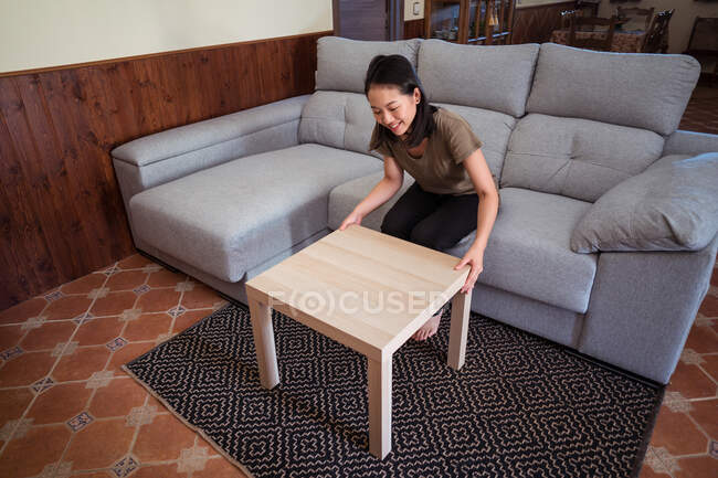 Cheerful young ethnic female mounting table on ornamental carpet against couch at home in daytime — Stock Photo