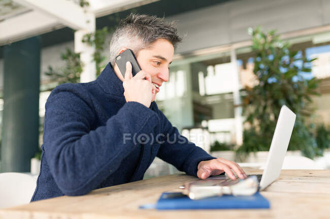 Вид сбоку молодого мужчины-фрилансера в повседневной одежде и очках, разговаривающего по мобильному телефону и смотрящего в камеру, сидящего за столом с ноутбуком и ноутбуком — стоковое фото