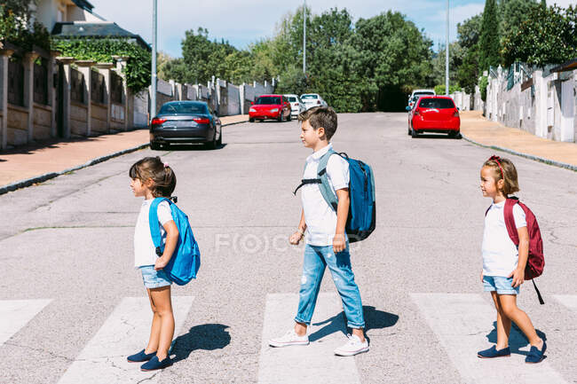 Вид сбоку на школьников с рюкзаками, стоящих на асфальтовой дороге и глядящих вперед в солнечный город — стоковое фото