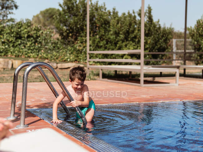 Позитивна дитина в шортах, що йдуть в басейн, розташований на задньому дворі в сонячний день влітку — стокове фото