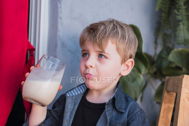 Niño mirando hacia otro lado mientras bebe bebidas de vidrio en vidrio mientras está sentado en un taburete hecho a mano - foto de stock