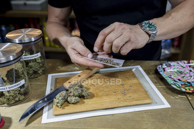 Crop maschio anonimo in orologio da polso con foglie di marijuana macinate asciutte su carta da sigarette sopra boccioli di fiori sul tagliere — Foto stock