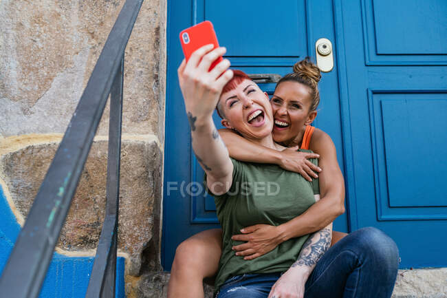 Mujeres homosexuales alegres con tatuajes tomando autorretrato en el teléfono celular contra la puerta de entrada en la ciudad - foto de stock