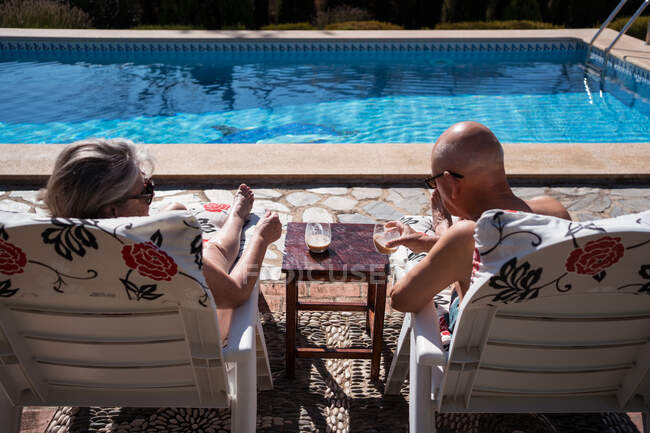 Вид ззаду на невпізнавану стару пару в купальнику, що п'є льодову каву під час охолодження на шезлонгах на задньому дворі — стокове фото