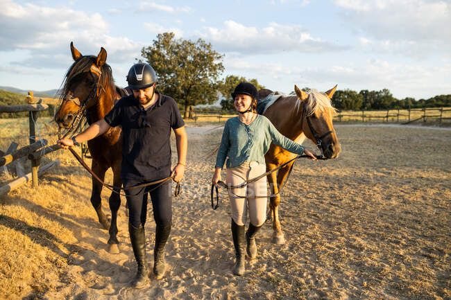 Smiling female and male trainer in protective helmets strolling with stallions on sandy terrain under cloudy sky — Stock Photo