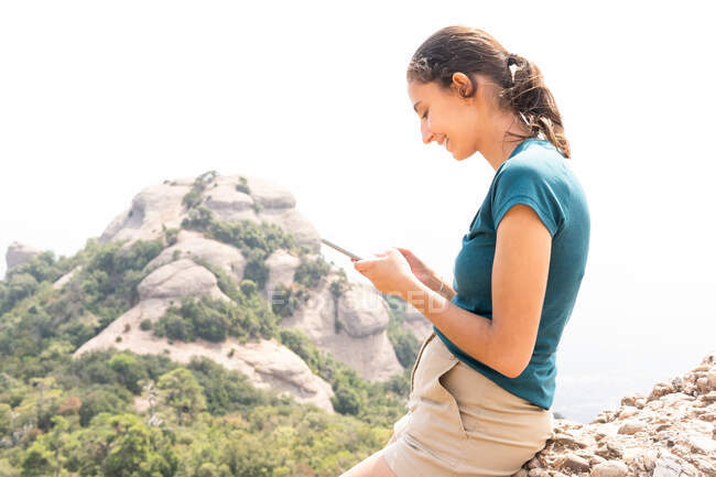 Vista lateral de mensajes de texto de turismo femenino positivo en el teléfono celular durante la excursión en las montañas de Montserrat en España - foto de stock