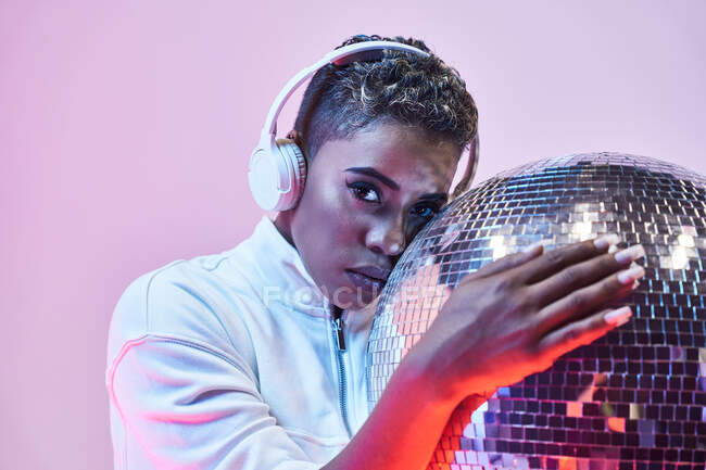 Cool ethnic woman in wireless headset with short hair and glitter ball looking at camera on violet background — Stock Photo