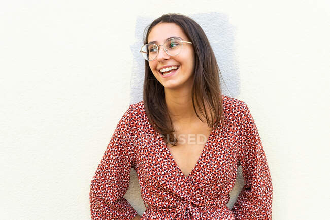 Positive Frau in stylischem Kleid und Brille, die in der Nähe von Lichtwänden steht und tagsüber wegschaut — Stockfoto