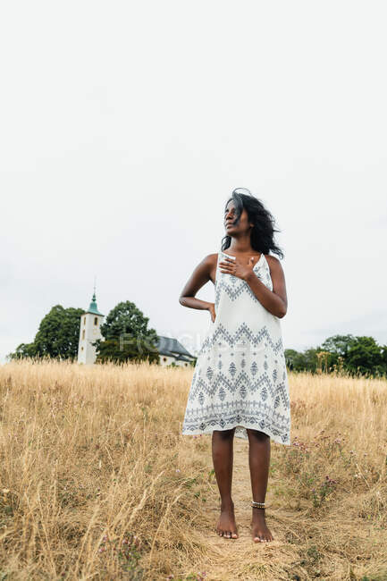 Босонога доросла етнічна жінка в сонячному одязі, торкаючись грудей, дивлячись на шлях проти каплиці під небом — стокове фото